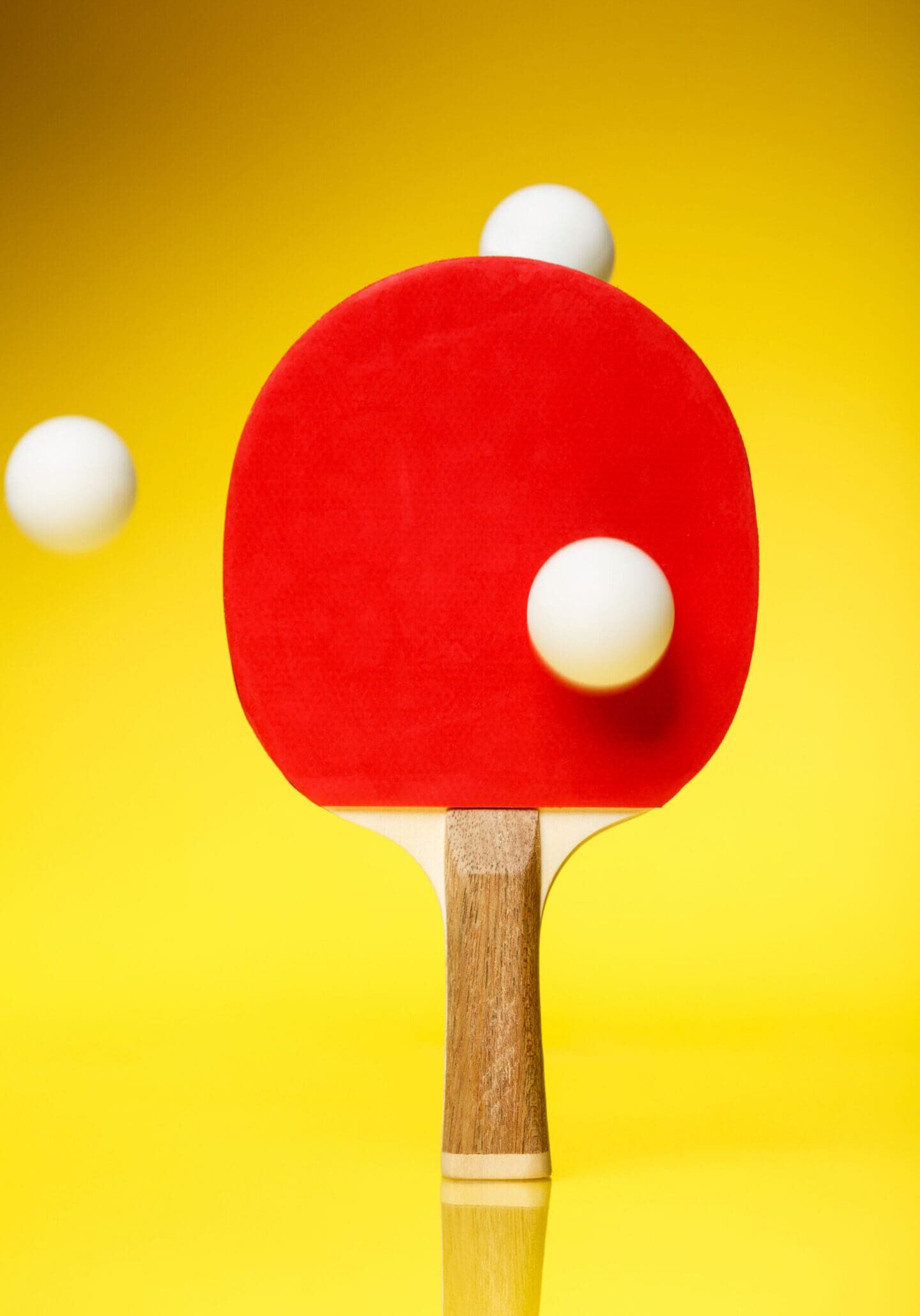 Red side of a ping pong racket with three white balls in the air, standing on a yellow background.